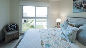 Quarto com uma cama grande, janela com vista para uma casa e gramado, uma cadeira de vime e uma mesa de cabeceira com abajur.