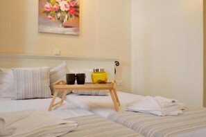 A hotel room with two beds, a tray table with cups and a yellow container, and a painting of flowers on the wall.