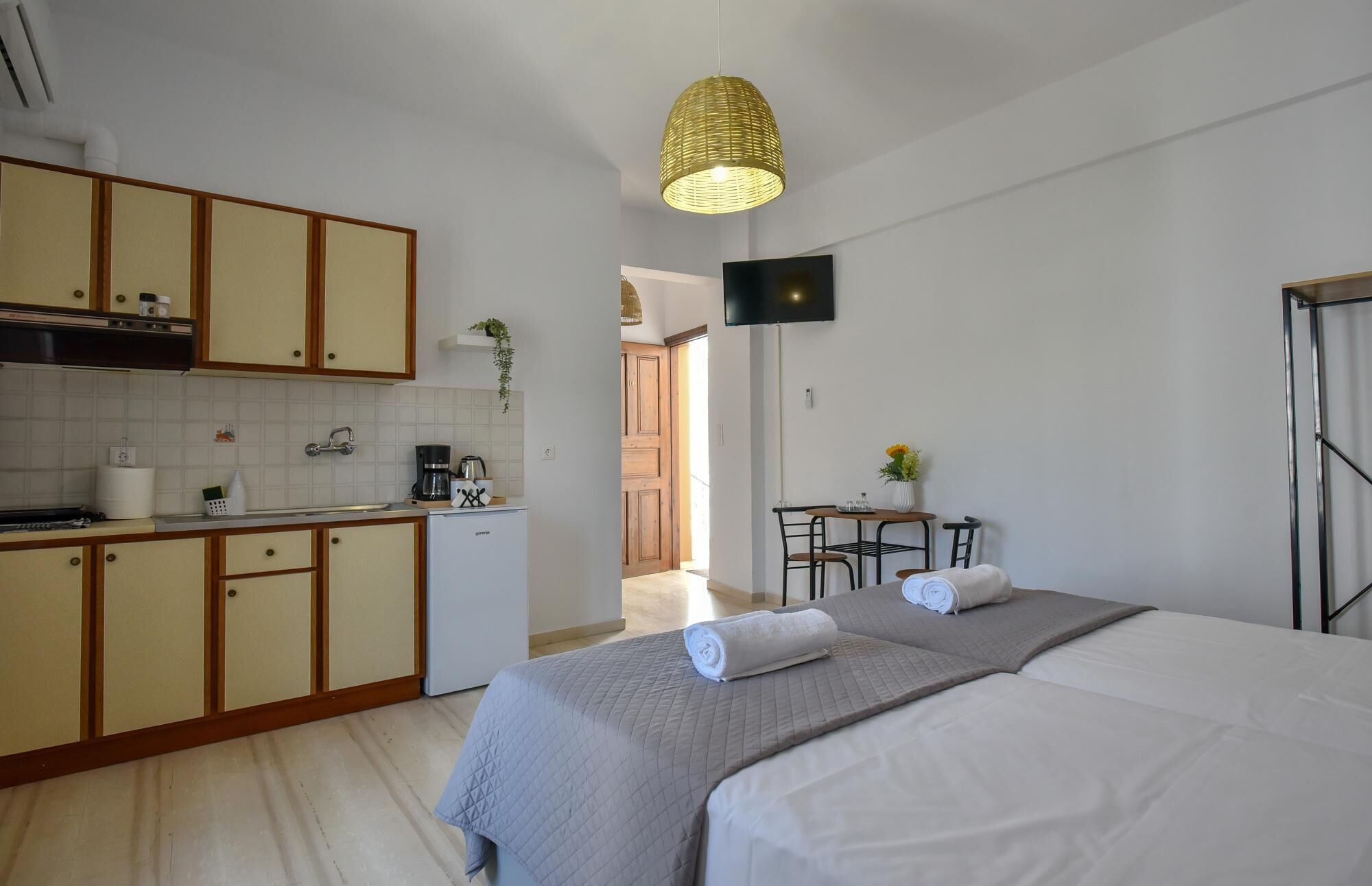 Ein kleines Hotelzimmer mit Küchenzeile, einem Bett mit zwei Kissen, einem Tisch mit Stühlen und einem an der Wand befestigten Fernseher.
