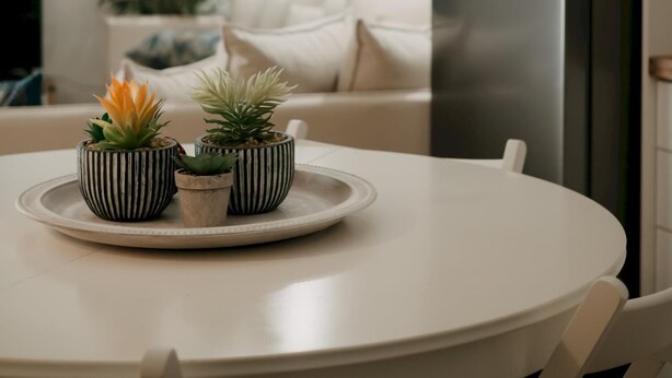 Une cuisine avec une table blanche, trois pots rayés contenant des plantes, un réfrigérateur et une fenêtre avec des stores.