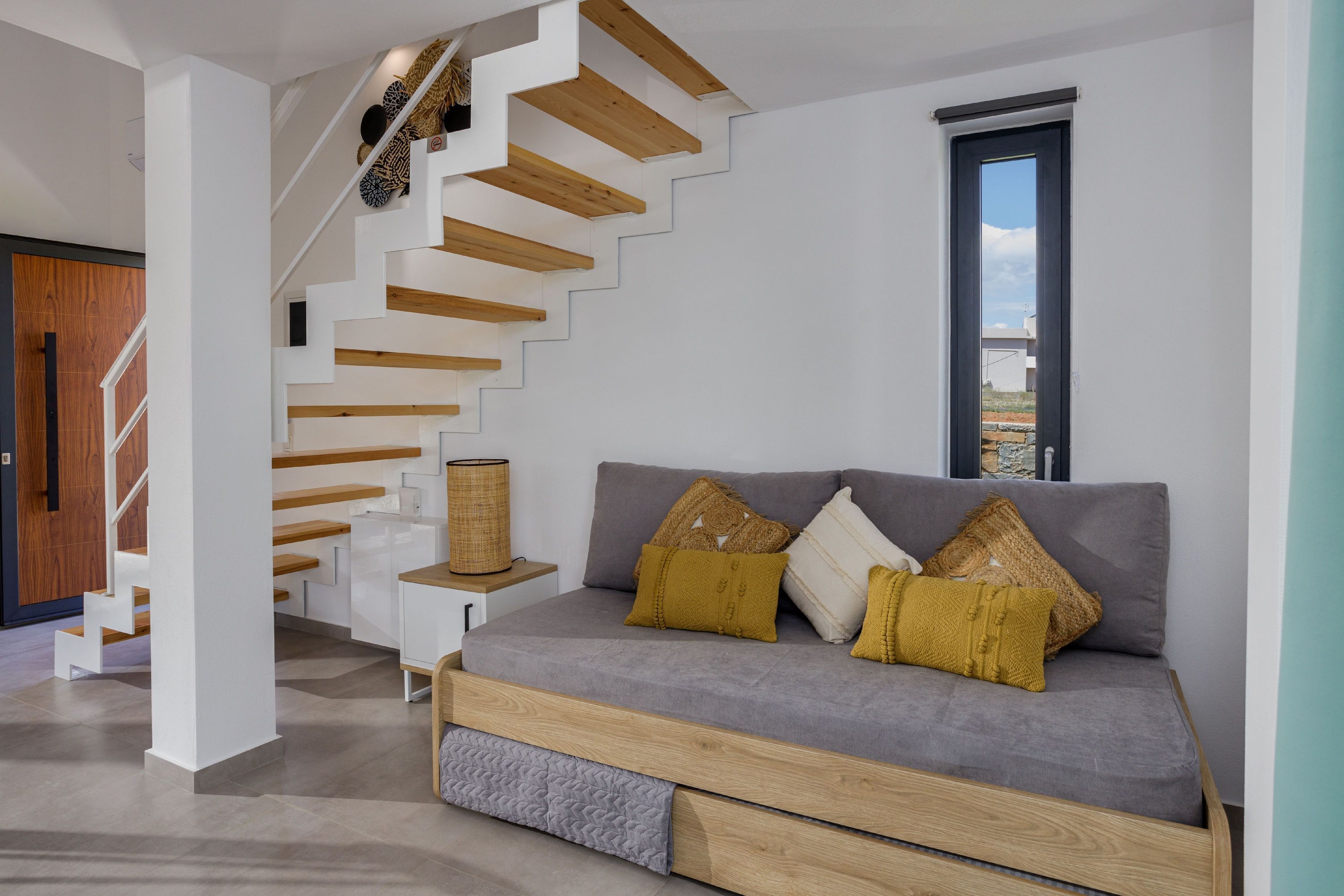 A modern interior with a staircase, a grey sofa with wooden legs, and a view through a window.