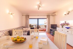 A modern living room with a sofa, dining table, and TV. There is a balcony with a view of the sea. - Sofia Beach Apartment by Acasadascasas (Mafra)