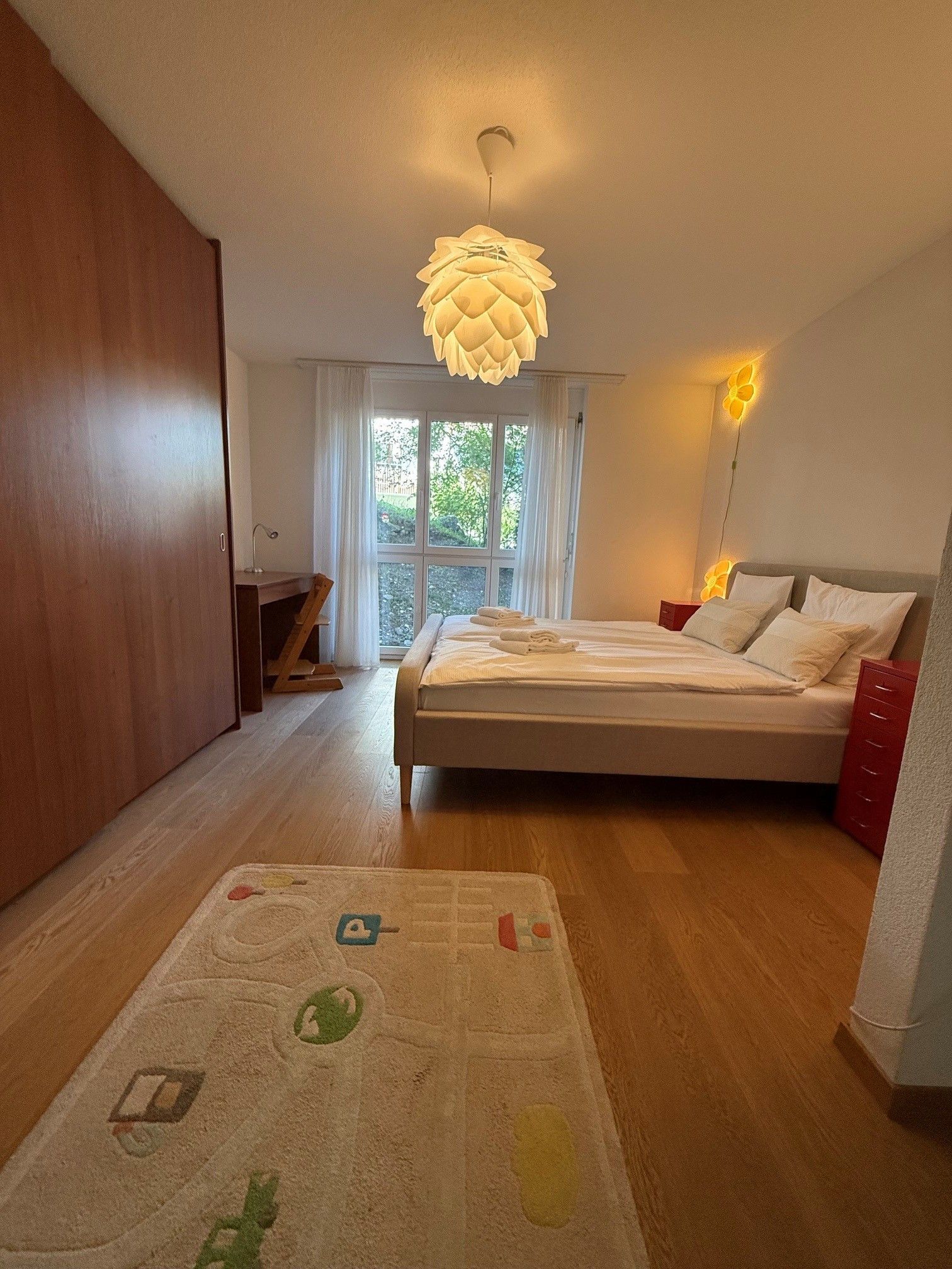 A bedroom with a bed, a desk, a chair, a window, and a hanging light fixture.