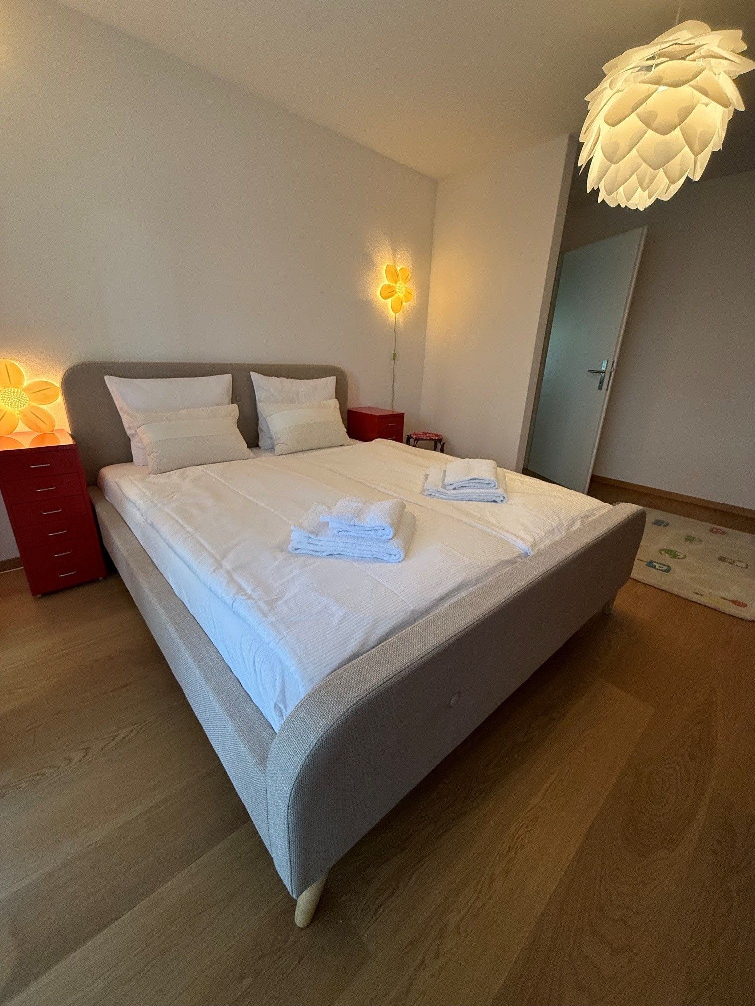 Un lit parfaitement fait, avec du linge de lit et des serviettes blanches, une tête de lit moderne et un luminaire mural original.