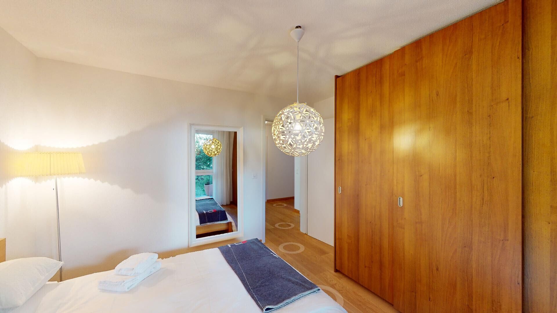 A bedroom with a bed, a wooden wardrobe, a hanging light fixture, and a view of a garden through an open door.