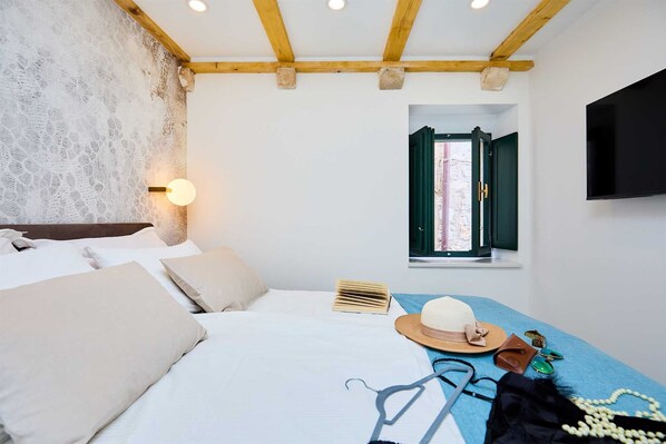 A bedroom with a bed, a wall-mounted light, a window with green shutters, and a TV on the wall.