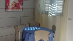 A dining area with a round table covered by a blue tablecloth, surrounded by four chairs. A framed artwork is on the wall, and there is a window with sheer curtains. - DUBROVNIK HAUS suites Standard studio apartment (Dubrovnik)