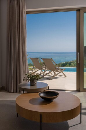 A modern room with a round wooden table, a bowl, and two chairs. A large window offers a view of the sea and a balcony with two chairs. - Villa Onyx - With Private Heated Pool (Rethymno)