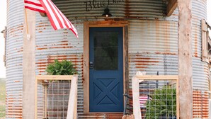 Un bâtiment rustique à l’extérieur en tôle ondulée, doté d’un porche en bois, d’une porte bleue et d’un drapeau américain.
