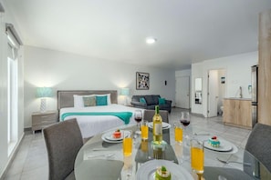 Quarto de hotel moderno com uma cama, um sofá, uma mesa de jantar posta para dois e uma cozinha.