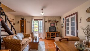 A cozy living room with a sofa, a coffee table, a TV stand, and a dining table. There is a staircase with a railing on the left, and a door leading outside. - Chalet do Monte Porto Moniz by Madeira Sun Travel (Porto Moniz)