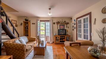 A cozy living room with a sofa, a coffee table, a TV stand, and a dining table. There is a staircase with a railing on the left, and a door leading outside.