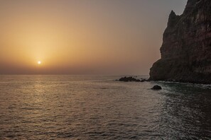 A coastal sunset with a rocky cliff and calm sea. - Madalena Sunset by Homie (Ponta do Sol)