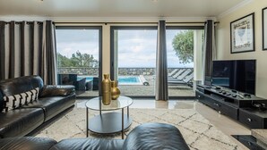 Ein modernes Wohnzimmer mit einer Ledersofa, einem Glastisch und einem Flachbildfernseher. Große Fenster bieten Blick auf einen Pool und Sitzgelegenheiten im Freien.