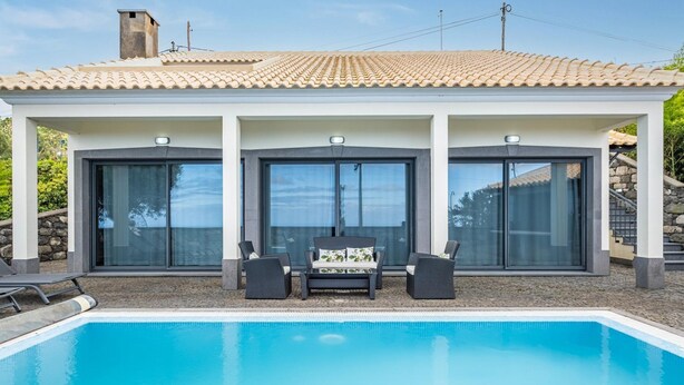Ein moderner Poolbereich mit einem klaren, blauen Pool, einer überdachten Lounge-Ecke mit zwei Stühlen und einem Tisch, sowie einem Gebäude mit einem bepflasterten Dach und großen Glasfenstern.
