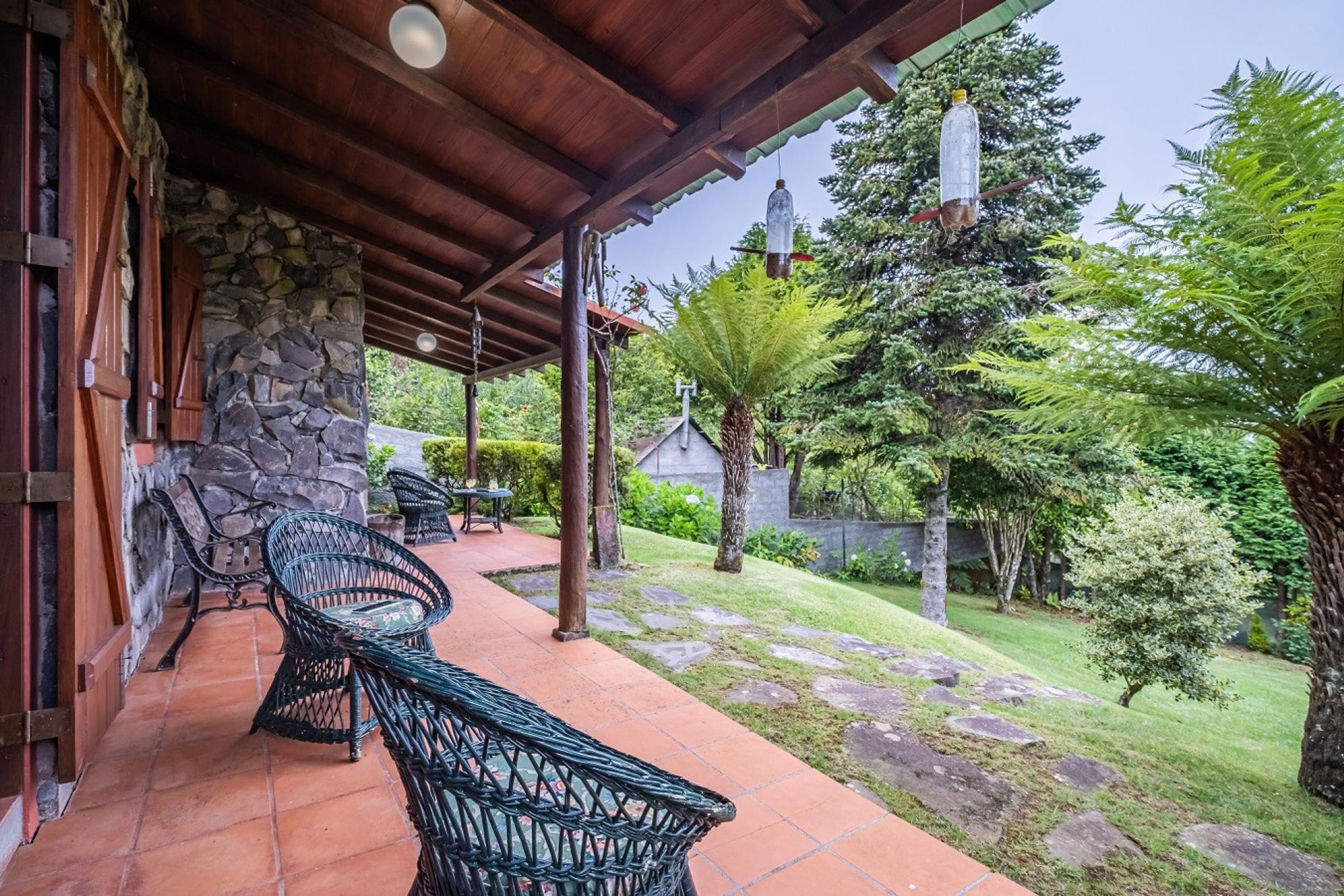 A covered patio with stone walls, outdoor furniture, and a garden with trees and a lawn.
