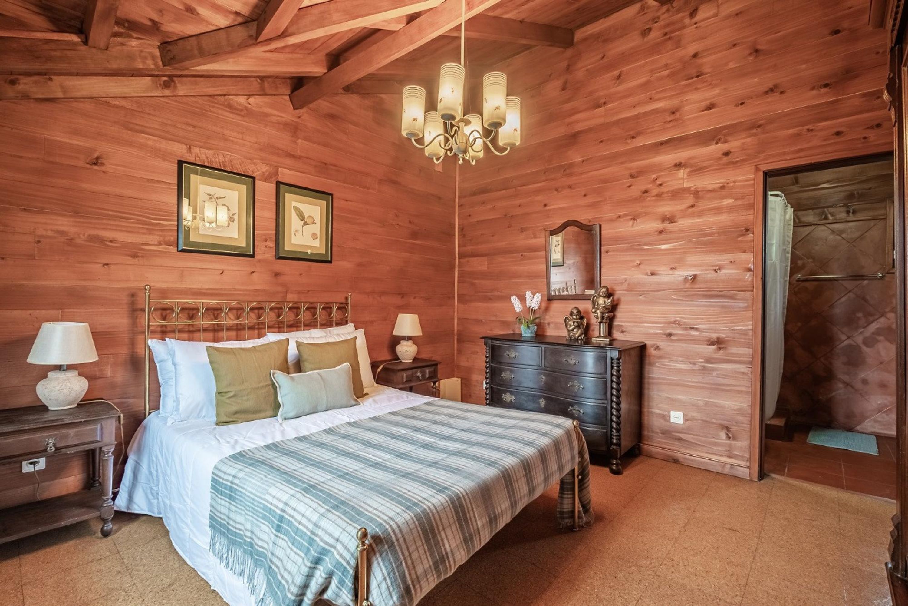 A wooden-paneled bedroom with a bed, two bedside tables, a dresser, a chandelier, and framed pictures on the wall.