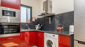 A compact kitchen with red cabinets, a built-in oven, a washing machine, and a coffee maker. - Ocean Panorama Apartment 2 by Homie (Calheta)