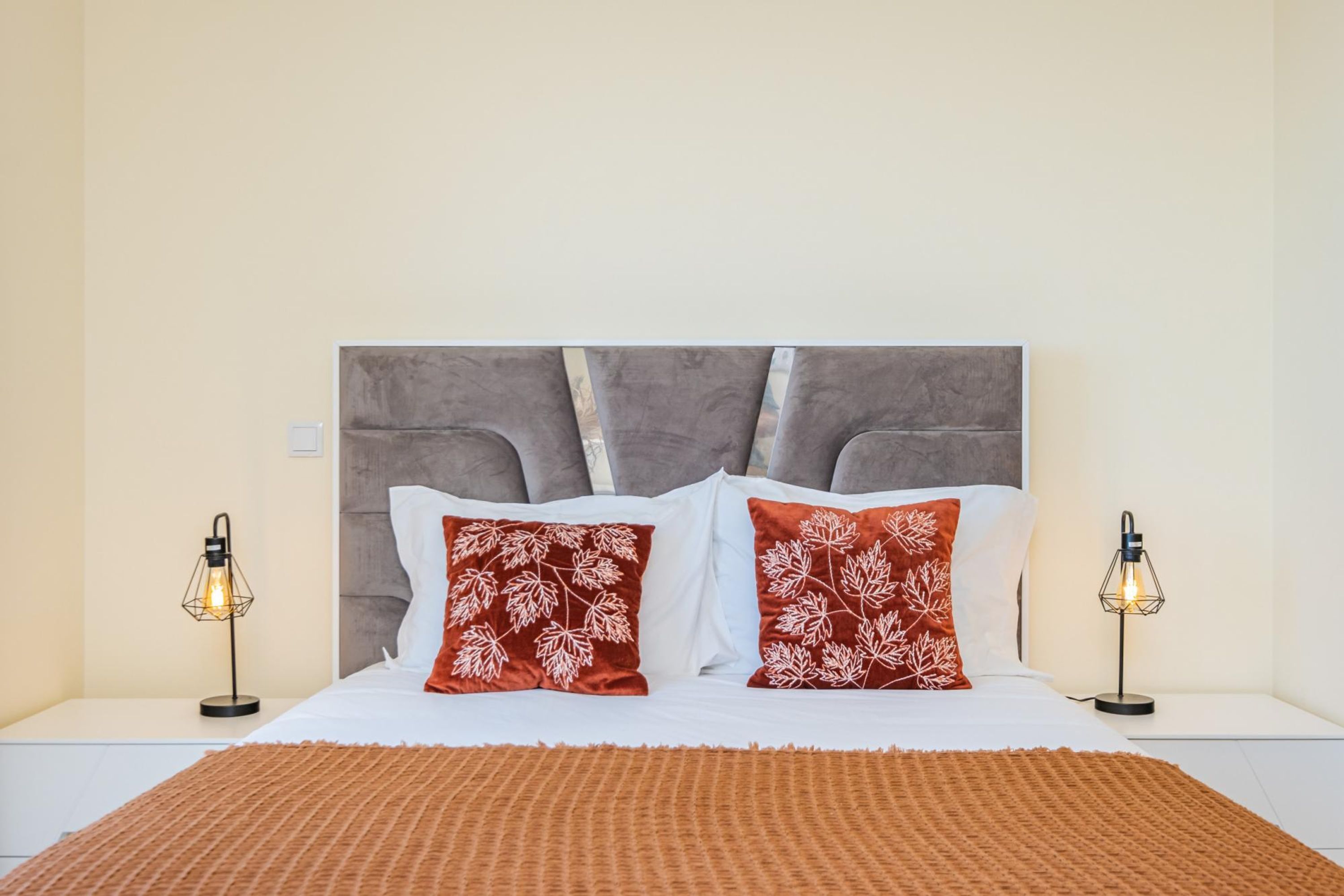 A neatly made bed with a grey headboard, two red pillows with white leaf patterns, and two bedside lamps on either side.