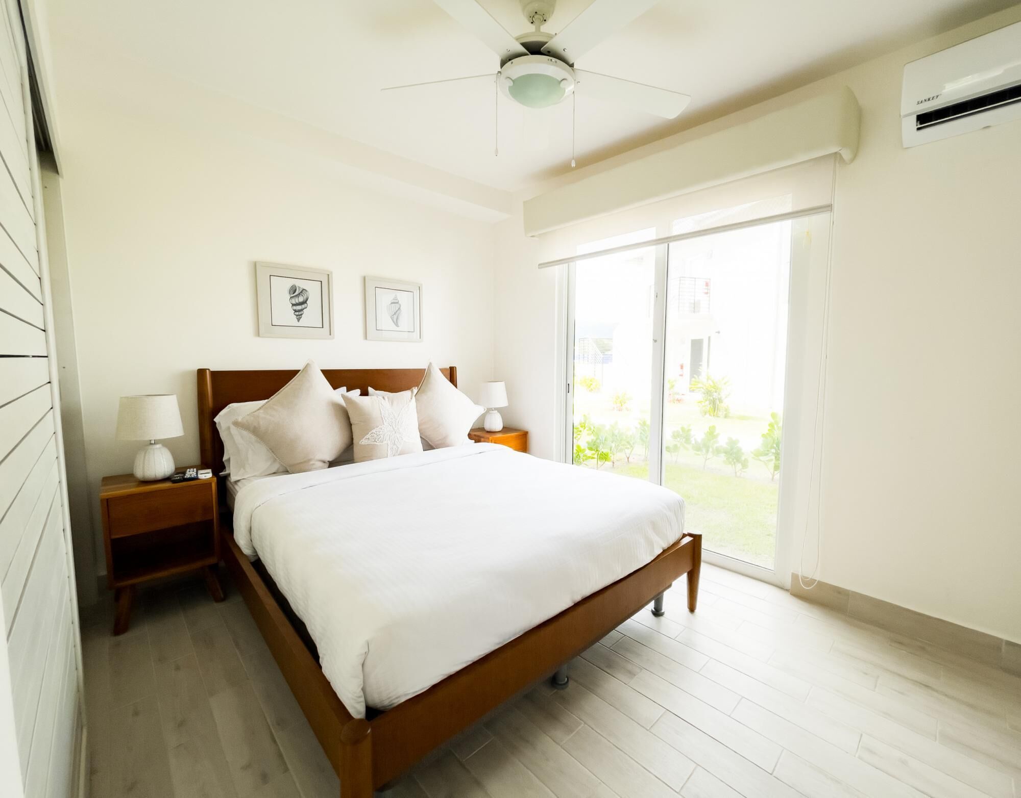 A bedroom with a wooden bed, white bedding, two bedside tables with lamps, a ceiling fan, and a large window with a view of greenery.