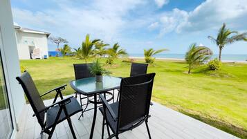 Un patio con una mesa de vidrio y cuatro sillas, con vista a un área verde con palmeras y una playa.