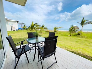 Une terrasse dotée d’une table en verre et de quatre chaises, offrant une vue sur un espace verdoyant agrémenté de palmiers et d’une plage.