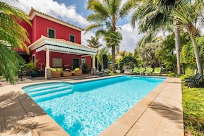 Una piscina con borde de baldosas, rodeada de palmeras y un edificio rojo con porche cubierto.