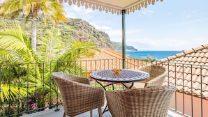 A balcony with wicker chairs and a table, overlooking a coastal landscape with palm trees and a view of the sea. - Quinta da Tia Briosa Casa da M e II by Homie (Ponta do Sol)