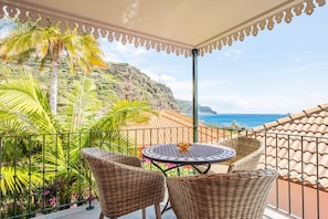 Un balcón con sillas de mimbre y una mesa, con vistas a un paisaje costero con palmeras y al mar.
