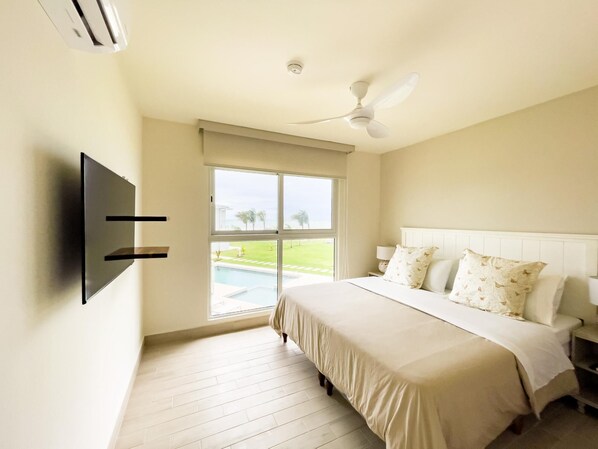 A bedroom with a bed, a ceiling fan, a TV mounted on the wall, and a window with a view of a swimming pool. - Residences C2-1d in Chame (Punta Chame)