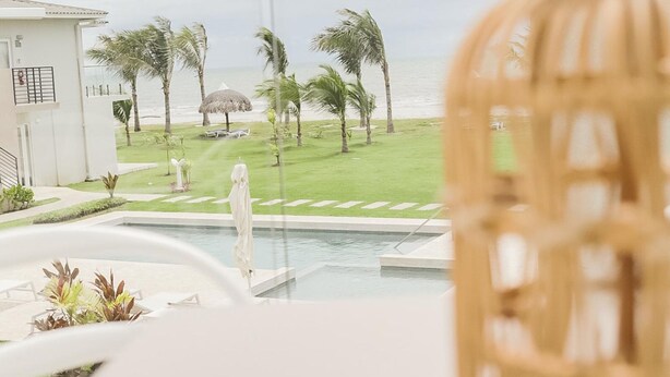 Blick vom Balkon auf einen Pool, mit einem Reetdachschirm und einem Strand im Hintergrund.