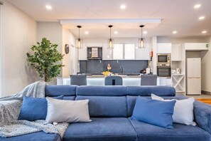 Een moderne woonkamer met een blauwe bank, een salontafel en een grote plant. Op de achtergrond een keuken met witte kasten, een magnetron en een koelkast.