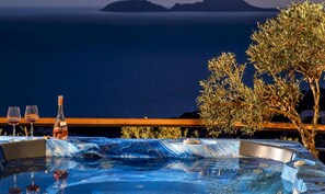 Un bain à remous avec vue sur la mer et les montagnes, deux verres à vin et une bouteille de vin posés sur une table.
