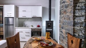 A modern kitchen with white cabinets, a stainless steel refrigerator, a black wood-burning stove, and a wooden dining table with plates of food. - Elena Junior Deluxe Suite by Estia (Agios Vasileios)