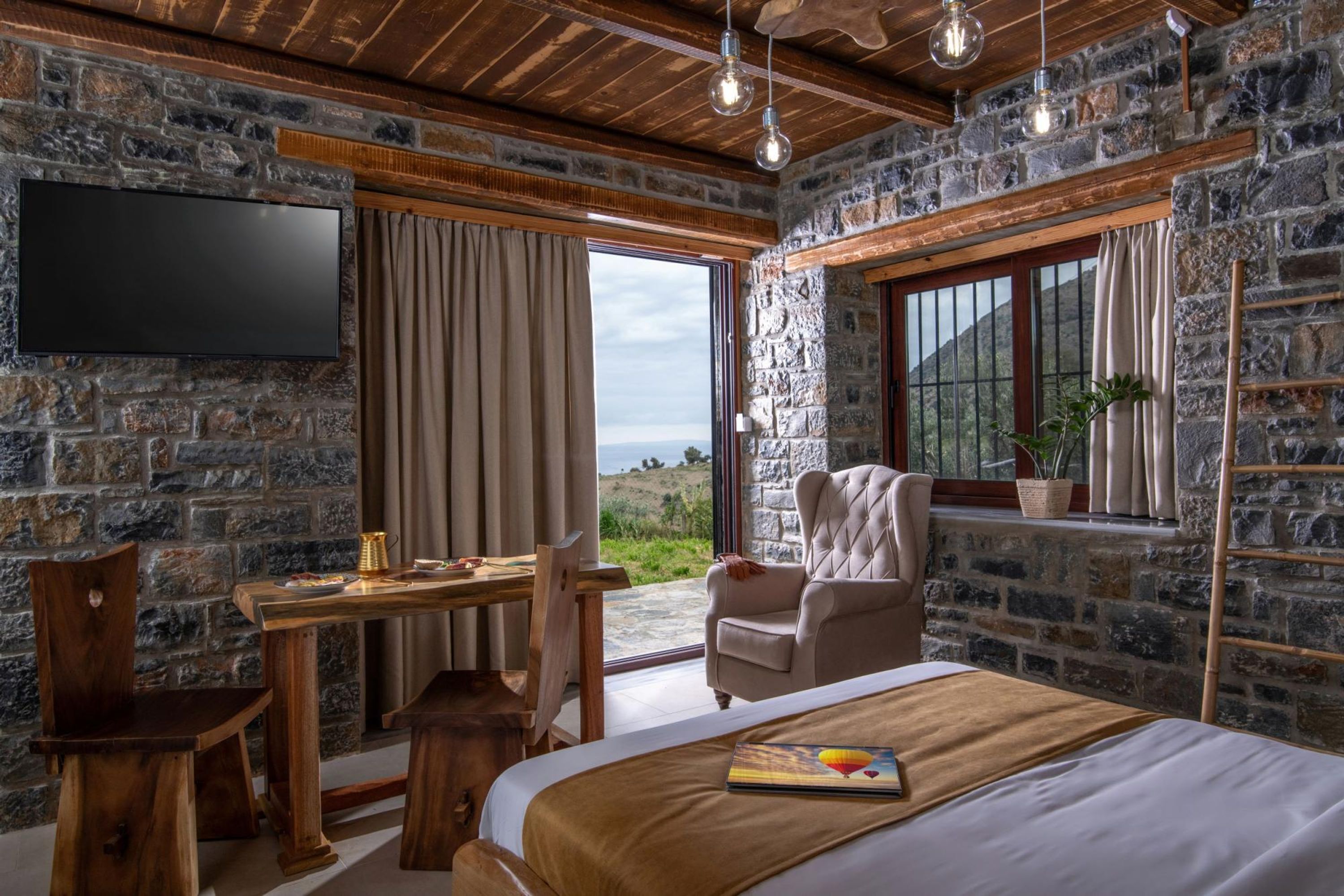 A room with stone walls, a wooden ceiling, a bed with a brown bedspread, a desk with a chair, a television mounted on the wall, and a window with curtains.