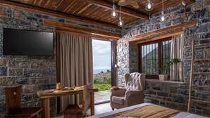A room with stone walls, a wooden ceiling, a bed with a brown bedspread, a desk with a chair, a television mounted on the wall, and a window with curtains. - Elena Junior Deluxe Suite by Estia (Agios Vasileios)