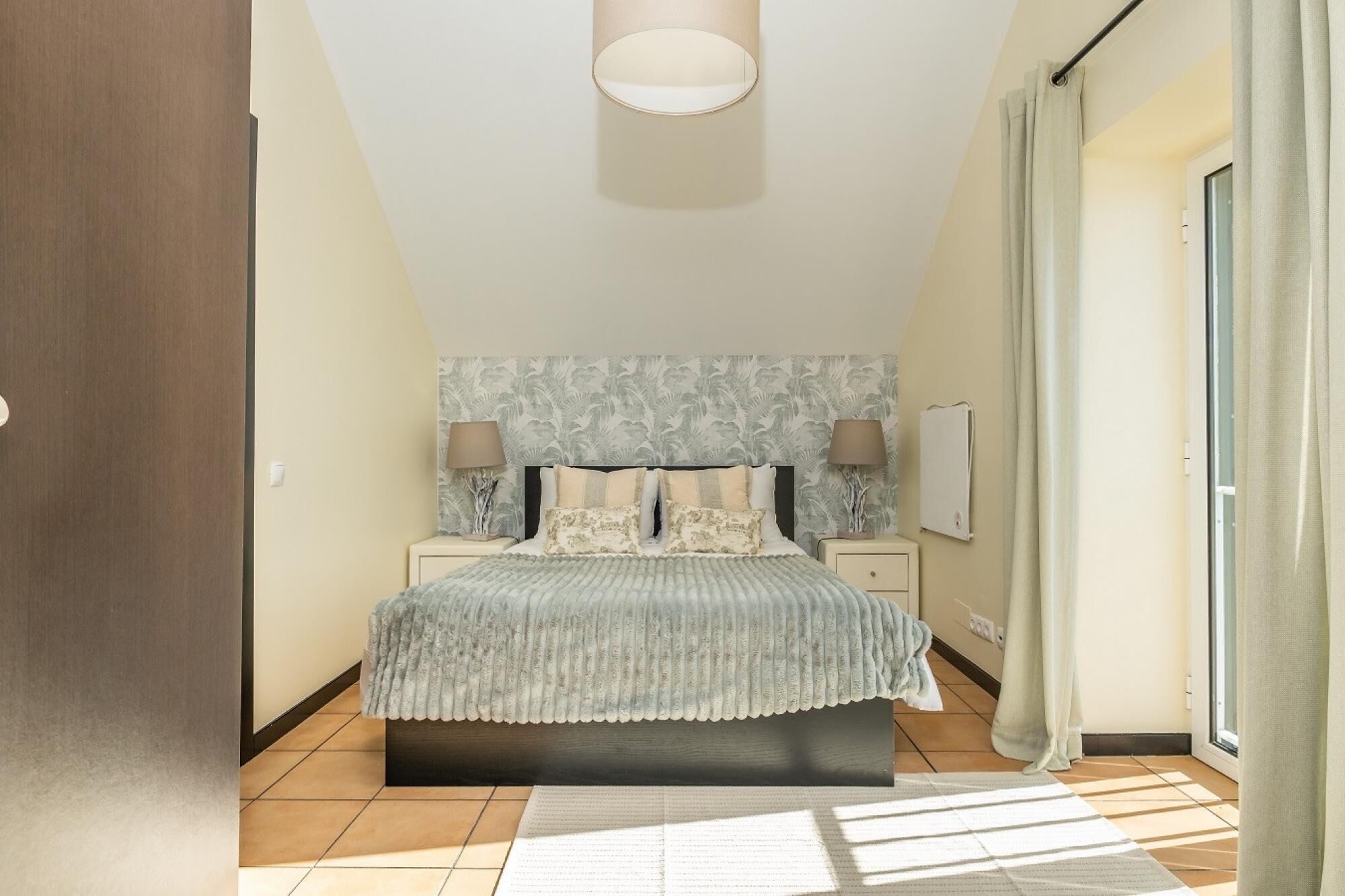 A bedroom with a bed, nightstands, a wall-mounted lamp, and a window with curtains.