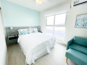 A bedroom with a bed, a nightstand, a teal armchair, and a window with a view. - Residences C14-1d in Chame (Punta Chame)