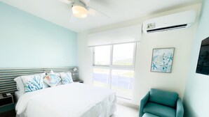 A bedroom with a bed, a teal armchair, a nightstand, a window, and an air conditioning unit. - Residences C14-1d in Chame (Punta Chame)