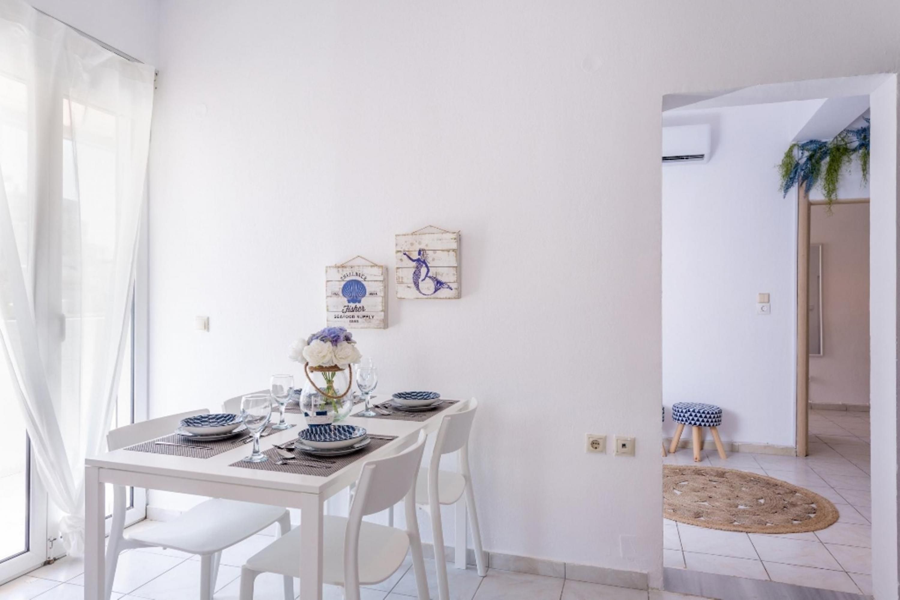 A dining area with a white table set for four, two wall-mounted decorations, and a view into another room with a plant.