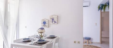 A dining area with a white table set for four, two wall-mounted decorations, and a view into another room with a plant.