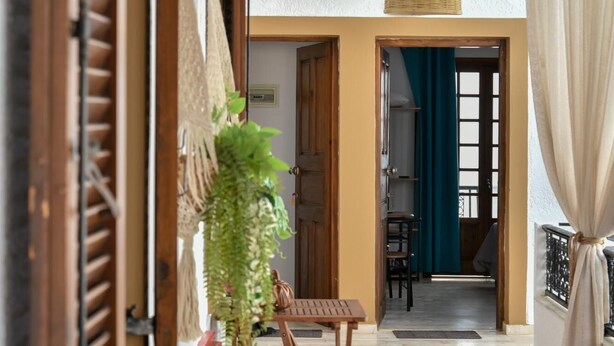A spacious living area with a wooden table, chairs, a hanging light fixture, and a view into another room with a blue curtain. - Eva Studios 10 in Agia Pelagia (Malevizi)