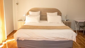 Ein ordentlich bezogenes Bett mit Holz-Kopfteil, ein Nachttisch mit Lampe und ein Sessel.