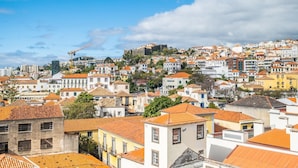Un paisaje urbano con una fortaleza imponente en una colina, edificios residenciales densos con tejados de tejas anaranjadas, y un cielo azul despejado.