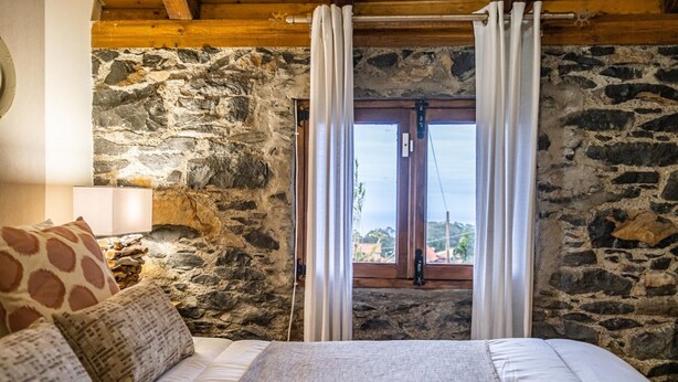 A bedroom with a stone wall, a wooden ceiling, a bed with white bedding, and a window with curtains. - King Shelter by Homie (Porto Moniz)