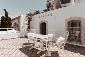Une terrasse extérieure blanche, avec une table et des chaises, une arche décorative et une fenêtre à motifs géométriques.