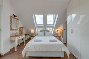 A bedroom with a large bed, two bedside tables, a dresser, and a wardrobe. There are skylights above the bed and a 'Welcome' sign on the wall.