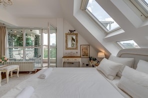 A bedroom with a large bed, a bedside table, a dresser, a mirror, and a window with a view of greenery and buildings.