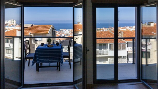 A balcony with a table set for two, overlooking a coastal town with red-roofed buildings and the sea. - Belle Vue Apartment by Acasadascasas (Mafra)