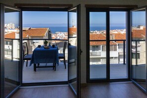 A balcony with a table set for two, overlooking a coastal town with red-roofed buildings and the sea. - Belle Vue Apartment by Acasadascasas (Mafra)